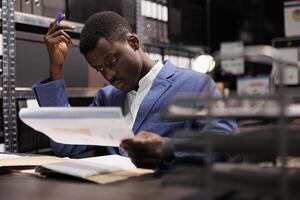 Corporate employee analyzing bookkeeping report, checking administrative record. African american businessman in formal suit searching for management document, working overtime in arhive room photo