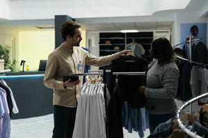 Clothing store employee showing jacket on hanger to customer, offering fashion advice. Shopping mall african american woman client and man worker choosing formal wear together photo