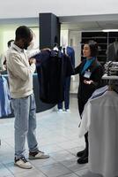 ropa Tienda asistente ofrecimiento cliente Consejo en elegir formal camisa. africano americano hombre y boutique trabajador participación formal vestir en percha, examinando estilo antes de haciendo compra foto