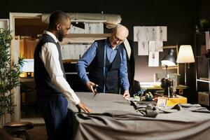 Meticulous elderly master tailor cutting fabric sheet for upcoming bespoke luxury fashion collection. Senior suitmaker showing african american apprentice expert craftmanship photo