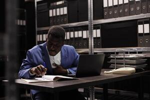 African american manager reading management research, working overtime at bureaucracy record in storage room. Businessman analyzing administrative files, searching for accountancy report photo