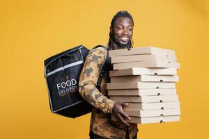 pizzería mensajero participación cajas lleno con pizza, entregando para llevar comida pedidos a clientes durante almuerzo tiempo. africano americano repartidor que lleva térmico mochila en estudio terminado antecedentes foto