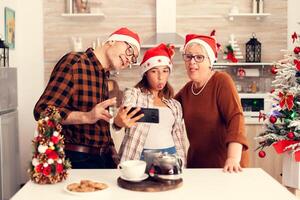 Niece celebrating christmas with grandparent making funny faces while taking selfies. Happy cheerful multi generation family using phone to take photo wearing santa hat doing hand gesture with christmas tree in the background.