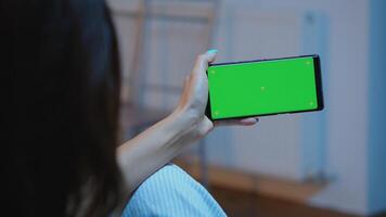 Lady looking at green screen smartphone while relaxing at home sitting on sofa. Woman holding a mobile phone with mockup template chroma key isolated cell phone display using techology internet photo