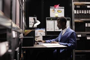 African american bookkeeper checking administrative files on laptop computer, working overtime in corporate depository. Businessman analyzing bureaucracy report in storage room photo