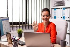 Businesswoman waving at notebook webcam during video conference with coworkers. Entrepreneur using internet connection for video meeting with coworkers looking at webcam. photo