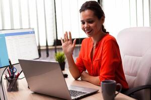 Businesswoman executive in video conference wave at team looking at camera. Entrepreneur using internet connection for video meeting with coworkers looking at webcam. photo