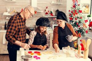 Family using shape cutter for dough on christmas day waring apron and bonette. Happy cheerful joyfull teenage girl helping senior woman preparing sweet cookies to celebrate winter holidays. photo