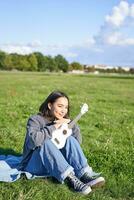 romántico asiático niña sentado con ukelele guitarra en parque y sonriente, relajante después universidad, disfrutando día apagado en Fresco aire foto