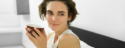 Close up of sensual young woman, looking aside and smiling, lying in bed, drinking morning cup of coffee, brown mug with drink photo