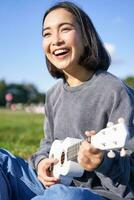 Vertical shot of smiling asian girl singing and laughing, playing ukulele, learn how to play instrument, sitting outdoors in park photo