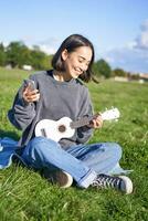 Vertical shot of smiling korean girl sitting in park with ukulele and smartphone, using mobile app to tune instrument, looking for chords while playing photo