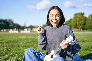 retrato de emocionado asiático chica, sentado con ukelele en parque en verde césped, mirando asombrado y sorprendido con bueno Noticias foto