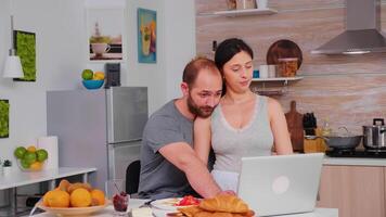 Happy couple surfing on internet using laptop during breakfast in kitchen. Married husband and wife in pajamas using internet web online modern technology, smiling and happy in the morning. Reading news browsing internet photo