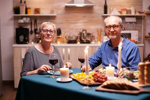 Joyful couple in kitchen looking at camera holding wine glasses looking at camera during dinner. Happy cheerful senior elderly couple dining together in the cozy kitchen, enjoying the meal. photo