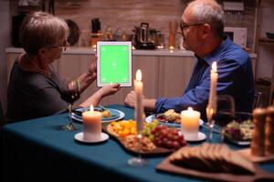 Old couple with green screen tablet pc in kitchen during festive dinner. Aged people looking at green screen template chroma key display sitting at the table in kitchen. photo