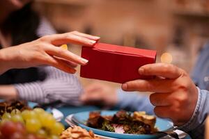 Close up of present box and couple having romantic dinner. Cheerful man dining with woman at home, enjoying the meal, celebrating their anniversary at candle lights. photo