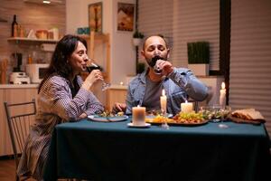 Pareja Bebiendo rojo vino teniendo relación celebracion. contento Pareja hablando, sentado a mesa en comida habitación, disfrutando el comida, celebrando su aniversario a hogar teniendo romántico tiempo. foto