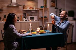 Boyfriend opening wine bottle. Happy couple talking, sitting at table in kitchen, enjoying the meal, celebrating their anniversary at home with healthy food photo