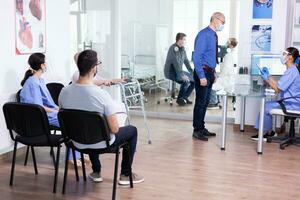 Senior man wearing sterile mask while answering questions from nurse in hopspital lobby during coronavirus and woman with walking frame sitting on chair. photo