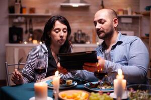 Pareja disfrutando romántico cena y utilizando tableta ordenador personal. . adultos sentado a el mesa en el cocina hojeada, buscando, , Internet, celebrando, aniversario, feliz., festivo disfrutar foto