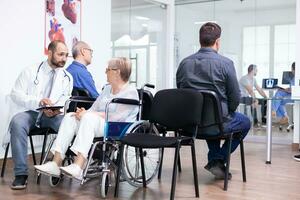 Doctor holding tablet pc with x-ray while explaining diagnosis to disabled elderly woman in wheelchair. Handicapped patient in hospital waiting area. Man in examination room. photo