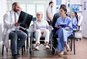 Doctor with stethoscope holding disabled senior woman radiography in wheelchair while talking with her in hospital waiting area. Patient asking about his appointment at clinic reception. photo