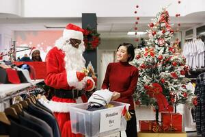 Asian woman helping with charity donations in clothing store, working with santa claus man to spread positivity during holiday season. Client and employee filling in donation box for kids. photo