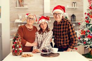 Cheerful multigeneration family wearing santa hat enjoying christmas with little girl smiling at camera. Happy loving grandparents celebrating winter holidays and relationship with granddaughter in home with x-mas decoration. photo