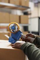 Cheerful employee using adhesive tape to pack cardboard box, preparing clients packages for delivery in warehouse. Storage room supervisor wearing industrial overall working at online orders. Close up photo