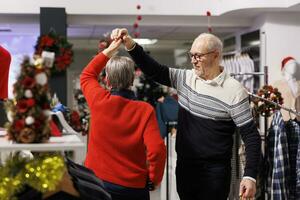 Cute couple dancing in retail store, moving around on ambience tunes and enjoying sweet moment. Elderly people showing true love and honest feelings to customers, doing waltz dance moves. photo