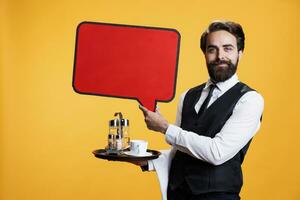 Skilled employee shows speech bubble icon in studio, working to create new marketing ad using red billboard. Young man butler in suit holding cardboard with empty isolated space on camera. photo
