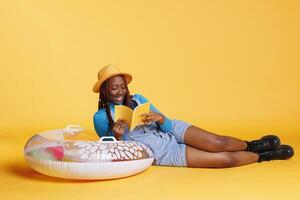 Female model reading book in studio, laying down on inflatable to enjoy lecture novel. Young adult feeling happy on camera, holding literature book and preparing to travel abroad. photo