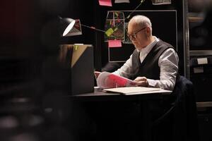 Elderly private detective sitting at desk table in dark room, working overtime at criminal case. Senior police officer analyzing missing person report, checking evidence documents photo