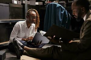 Detective and assistant sitting on floor surrounded by case files and working overtime in agency office. Two african american cops making research, searching crime solving solution photo