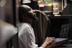 Tired police agent sitting with closed eyes, solving complex crime case in dark office. Exhausted private detective spying on suspect, watching cctv camera records on laptop photo