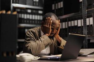 Exhausted african american investigator solving complex crime case in evening. Tired policeman holding hands on head while studying forensic expertise reports in office at night photo