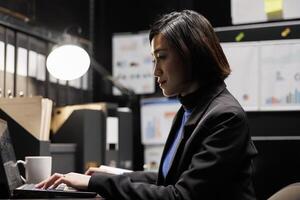 Professional businesswoman checking administrative analytical data in dark file room surrounded by accountancy statistic flowcharts. Asian worker in bureaucratic office filled with invoice folders photo