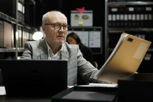 Senior private detective in criminal cases archive room revising criminal case file details. Elderly investigator surrounded by criminology folders on cabinet shelves in agency repository office photo