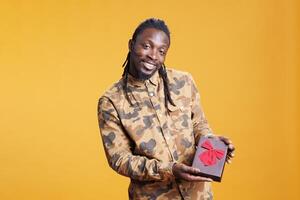 Confident smiling man presenting wrapped present box for girlfriend, posing for valentine s day in studio over yellow background. Handsome african american person celebrating love holiday. photo