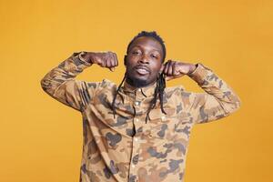 Athletic confident man flexing trained arm muscles in front of camera in studio over yellow background. African american young person enjoying sport exercices, working out at biceps strength. photo