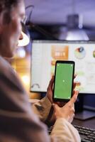 Entrepreneur looking at green screen mock up chroma key smartphone with isolated display, analyzing marketinf graphs report. Executive manager working late at night in startup office photo