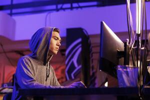 Asian hacker breaking law while doing data mining using malicious software. Young man in hood hacking into computer system while cracking password in abandoned warehouse at night photo