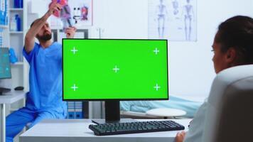 Doctor working on computer with green screen in hospital and assistant looking at x-ray. Desktop with replaceable mockup screen in medical clinic while doctor is checking patient radiography for diagnose. photo