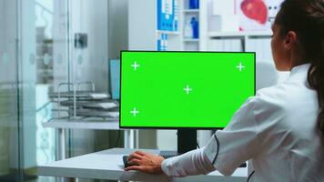Doctor sits at computer with blank green screen display in hospital cabinet and assistant in blue uniform holding x-ray. Medic in white coat working on monitor with chroma key in clinic cabinet to check patient diagnose. photo