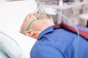 Sick elderly man infected with covid19 breathing through oxygen mask in private clinic. Patient in hospital room laying on medical bed with infected lung during coronavirus pandemic having respiratory problem. photo