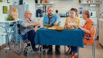 abuelo con invalidez en silla de ruedas y familia teniendo cena. dos contento parejas hablando y comiendo durante un gastrónomo comida, disfrutando hora a hogar sentado alrededor el mesa en el cocina. foto