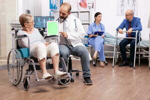 Handicapped senior woman sitting in wheelchair discussing with doctor holding tablet pc with green screen. Medical practitioner using device with chroma key during coonsultation of invalid senior woman. photo