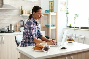 mujer mecanografía en ordenador portátil disfrutando verde té durante desayuno en cocina. trabajando desde hogar utilizando dispositivo con Internet tecnología, hojeada, buscando en artilugio en el Mañana. foto