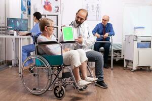 Hospital staff consulting handicapped senior woman sitting in wheelchair holding tablet pc with green screen. Medical practitioner using device with chroma key during coonsultation of invalid senior woman. photo